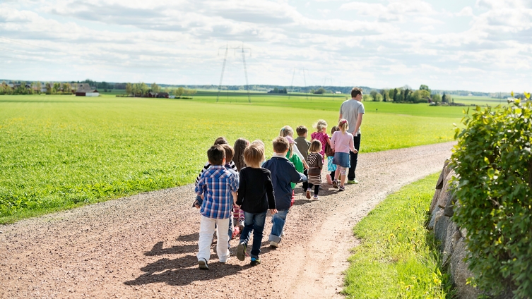 En förskolelärare och tolv barn går på en grusväg i ett sommarlandskap