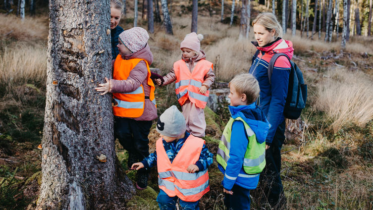 Två förskollärare ute vid en stor stubbe i skogen med fyra barn. Foto: Madeleine Wejlerud